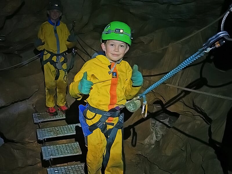 Deux enfants en tenue de spéléologie jaune et verte posant sur un pont suspendu dans une grotte.