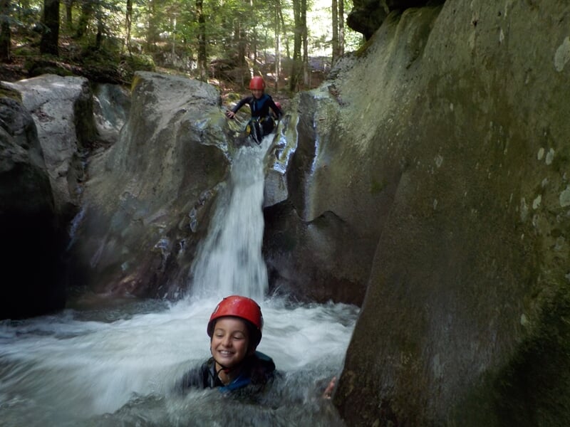 Enfant souriant dans une piscine naturelle au pied d'une petite cascade en pleine forêt lors d'une sortie canyoning.