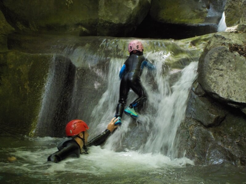Deux personnes en combinaison et casque rouge escaladant une cascade rocheuse avec un courant d'eau en canyoning.