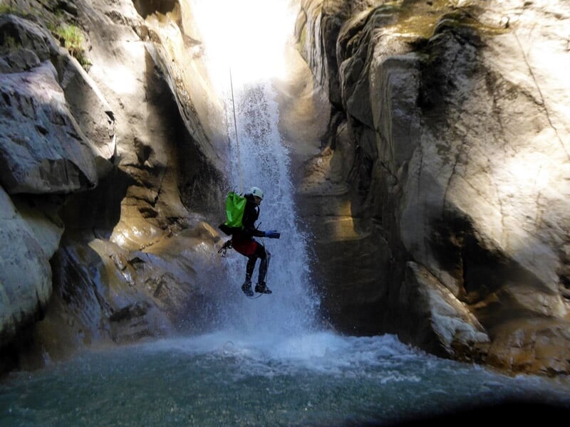 Personne suspendue en rappel devant une grande cascade dans un canyon rocheux éclairé par le soleil.