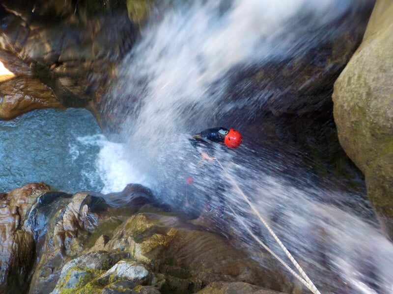 Personne en combinaison noire et casque rouge descendant une cascade en rappel dans un canyon avec eau turquoise.