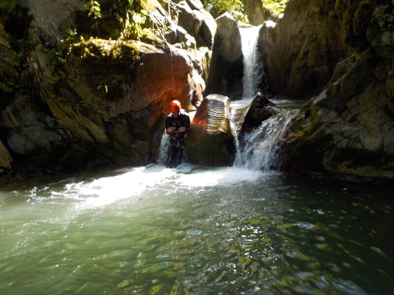 Personne assise dans une piscine naturelle au pied d'une cascade dans un canyon rocheux entouré de végétation.