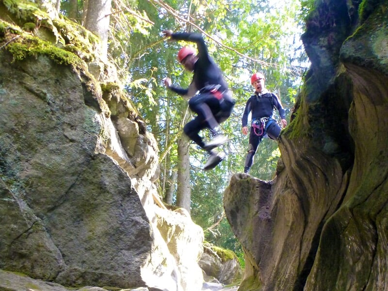 Personne sautant d'une falaise rocheuse dans un canyon, équipée d'un casque rouge et d'une combinaison noire.