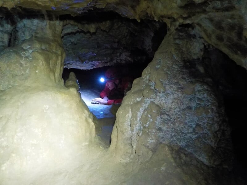 Explorateurs en combinaison rouge avec lampes frontales progressant dans une grotte étroite et rocheuse en spéléologie.