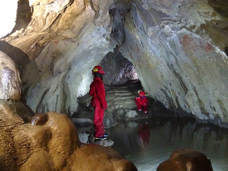 Deux spéléologues en combinaison rouge explorant une grande salle souterraine avec formations rocheuses et eau.