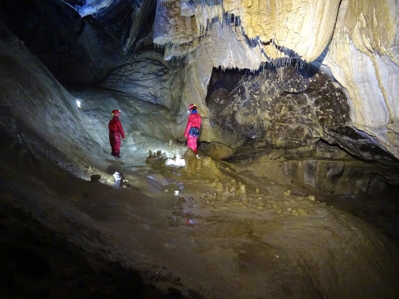 Deux personnes en tenue rouge avec casques et lampes frontales dans une grotte aux parois rocheuses et sol boueux.
