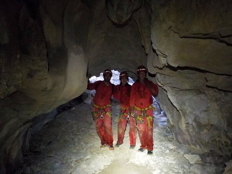 Trois spéléologues en combinaison rouge posant dans une galerie souterraine rocheuse éclairée par leurs lampes.