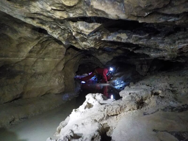 Spéléologue en combinaison rouge progressant à quatre pattes dans une galerie basse et étroite d'une grotte.