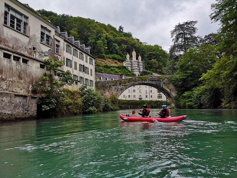 Deux personnes en kayak rouge pagayant sur une rivière calme avec un pont en pierre et des bâtiments anciens.