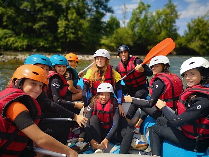 Groupe de jeunes en tenue de rafting avec casques et gilets de sauvetage souriant avant une descente en rivière.