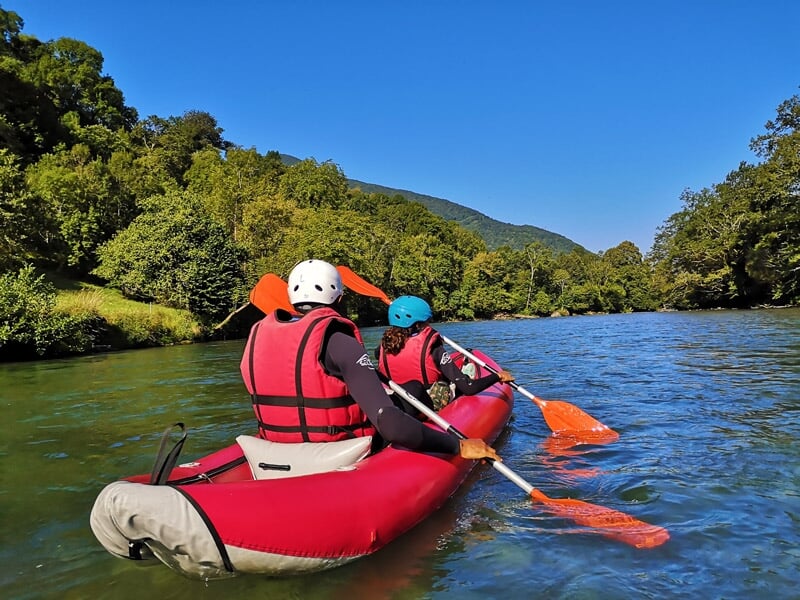 Deux personnes pagayant dans un kayak rouge sur une rivière calme bordée d'arbres verts et montagnes.