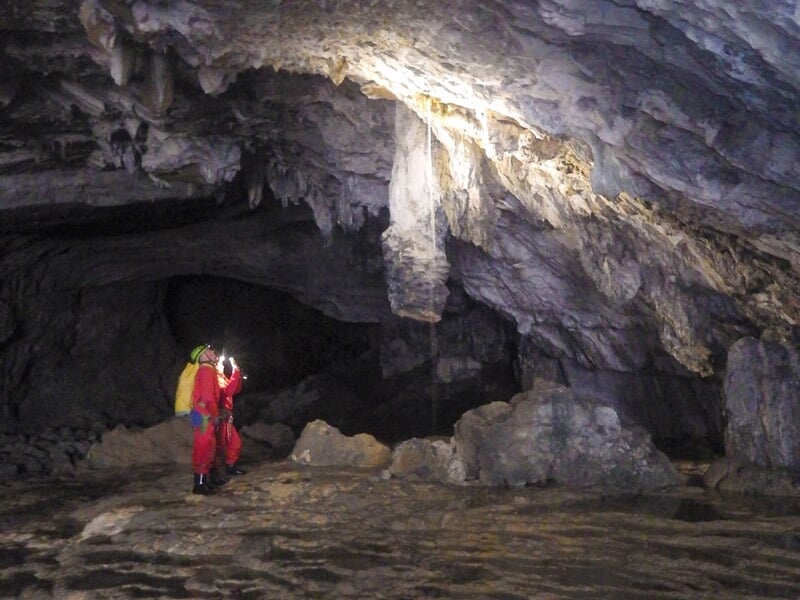 Spéléologues observant une stalactite imposante éclairée dans une grande cavité rocheuse, au sol irrégulier et humide