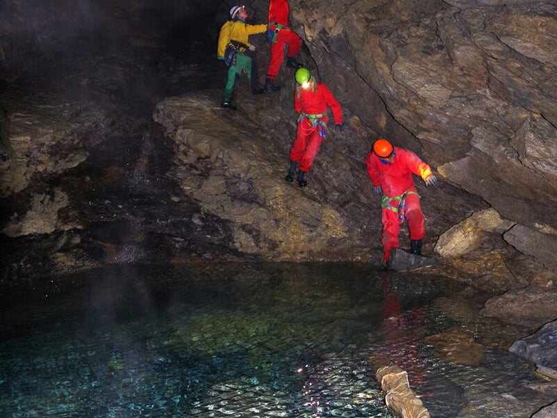 Participants à une exploration souterraine traversant un passage étroit au-dessus d’un bassin d’eau claire, équipés de casques et harnais