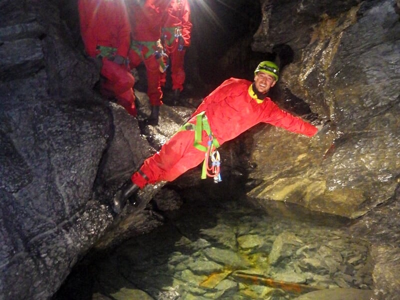 Spéléologue en combinaison rouge s’appuyant sur une paroi rocheuse au-dessus d’une petite rivière souterraine, éclairé par une lampe frontale