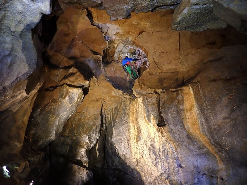 Spéléologue équipé descend dans une grotte rocheuse sombre avec un casque et une lampe frontale.