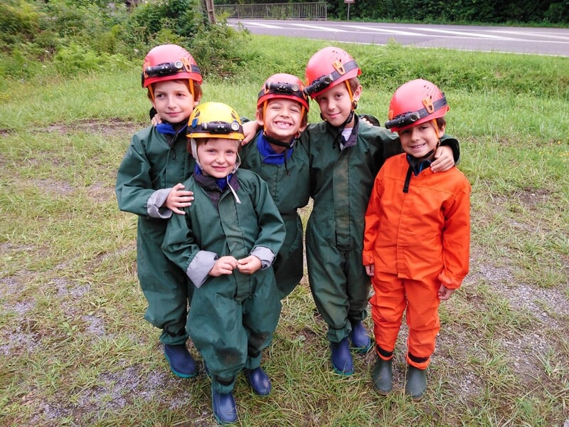 Cinq enfants en combinaison et casque rouge et orange posent souriants en extérieur avant une activité spéléologie.