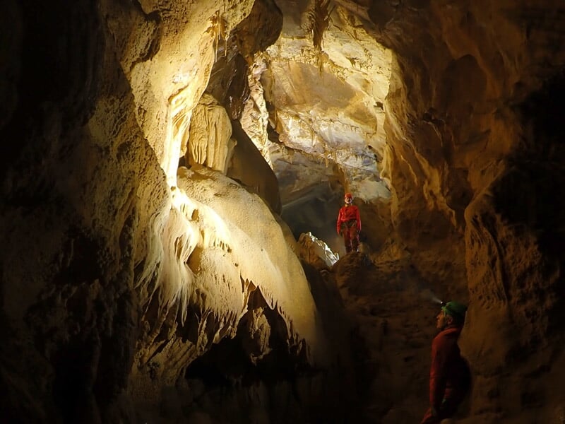 Personne en combinaison rouge et casque descend en rappel dans une grotte rocheuse éclairée par une lampe.