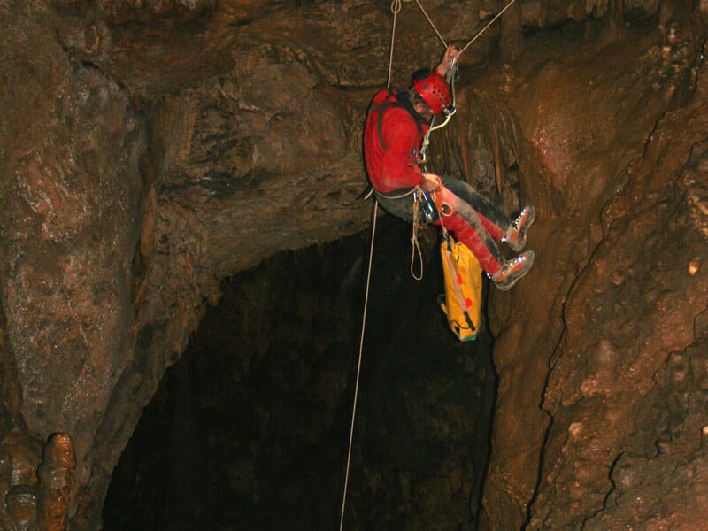 Deux spéléologues explorent une grande salle souterraine avec des stalactites et stalagmites éclairées.