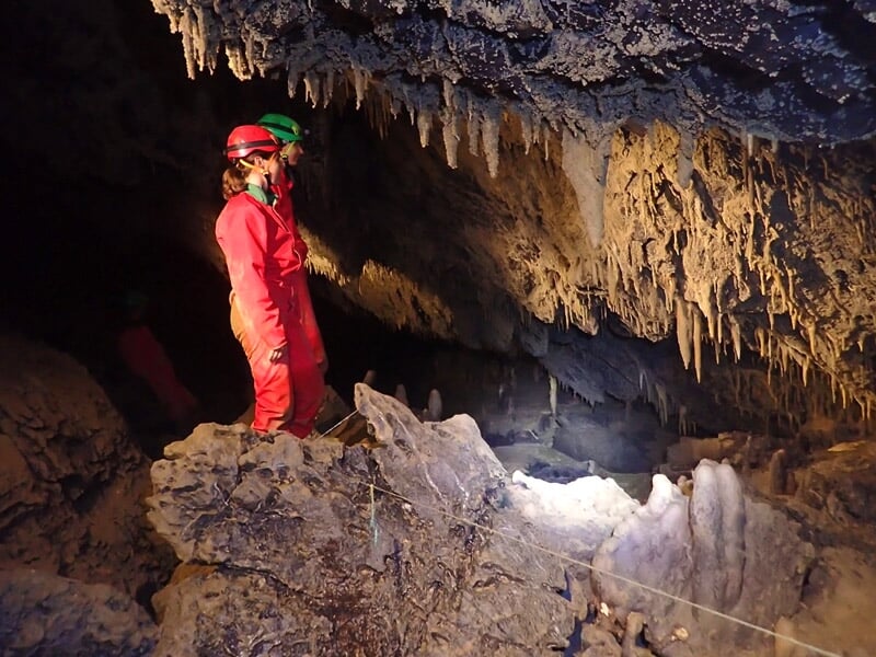 Deux spéléologues en combinaison colorée explorent une cavité rocheuse avec stalactites et sol irrégulier.