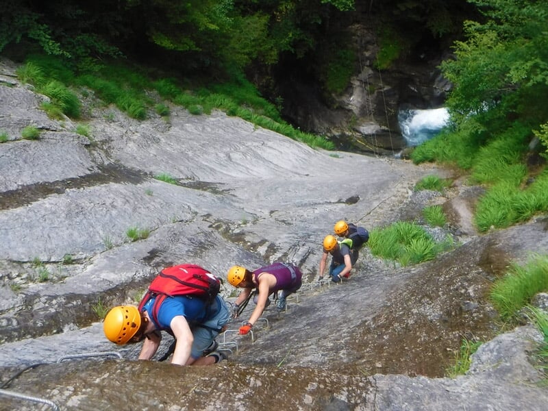 Trois personnes en casque et harnais escaladent une paroi rocheuse abrupte en via ferrata sous un ciel bleu.