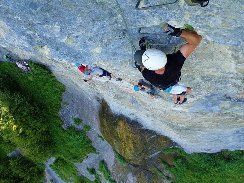 Personne en casque blanc et harnais grimpe une paroi rocheuse verticale en via ferrata avec vue sur la vallée.