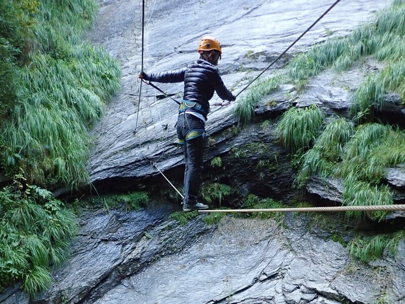 Personne en tenue d'escalade et casque blanc avance sur un pont suspendu en via ferrata au-dessus d'un ravin.