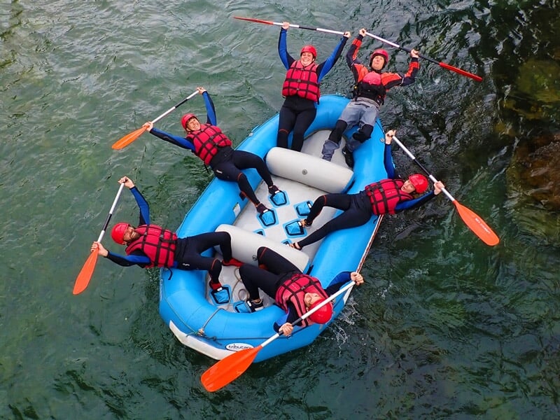 Personnes en raft bleu pagayant dans une rivière entourée d'arbres verts et rochers, activité sportive.