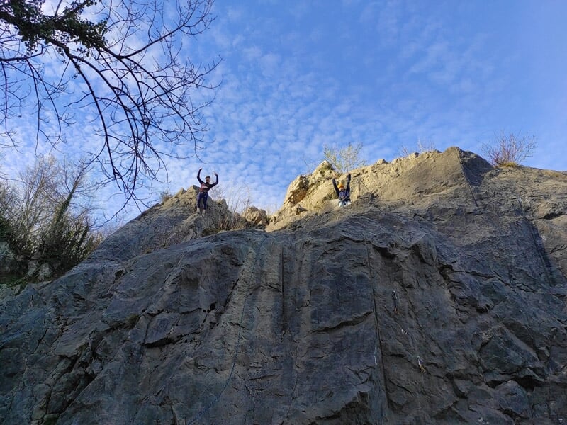 Grimpeur en casque jaune et harnais escalade une paroi rocheuse avec vue sur un paysage verdoyant.