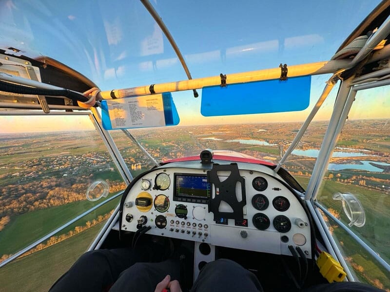 Vue intérieure d'un cockpit d'ULM en vol, surplombant un paysage champêtre au coucher du soleil, tableau de bord équipé d'instruments de navigation, vitres teintées en bleu