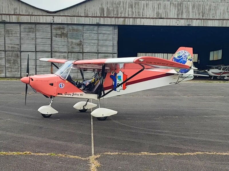 ULM rouge avec des accents blancs et une empreinte de globe sur l'empennage, stationné devant un hangar, slogan