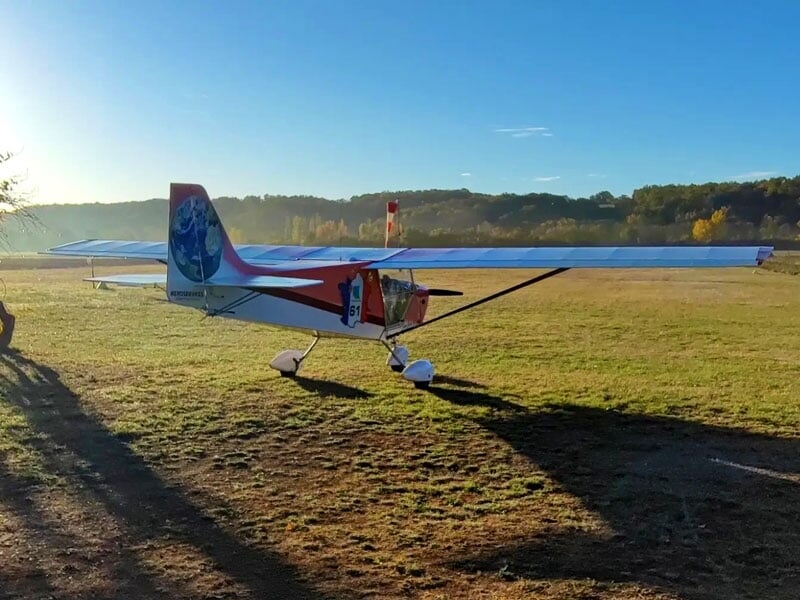 ULM rouge et blanc avec empennage arborant une empreinte de globe terrestre, stationné sur un terrain en herbe, ombre allongée au sol, soleil bas