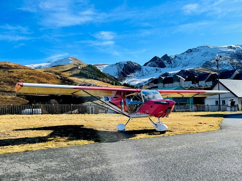 ULM rouge et blanc garé en plein air, montagnes enneigées en arrière-plan, ciel bleu, village alpin adjacent