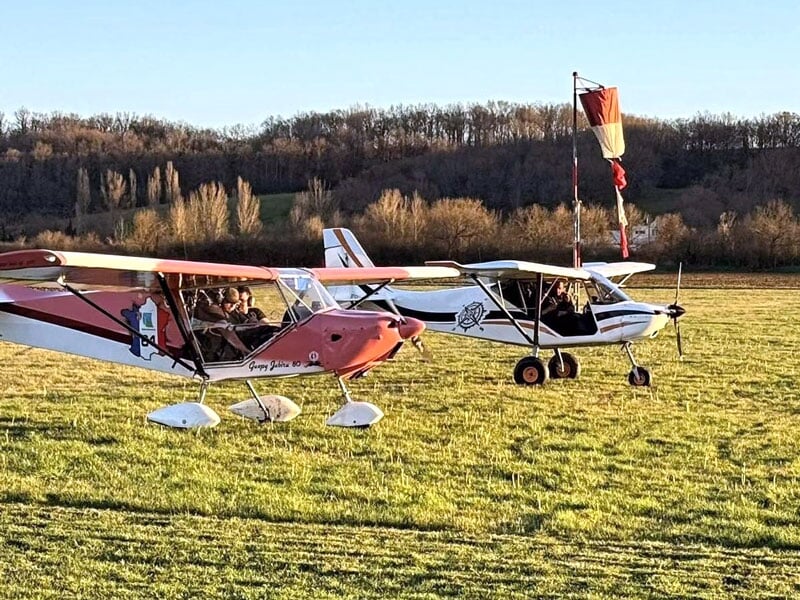 Deux ULMs stationnés sur une piste herbeuse en fin d'après-midi, le premier de couleur rouge avec un motif de carte à jouer, le second blanc, ciel clair