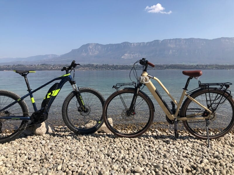 Deux vélos électriques posés sur un sol caillouteux au bord d'un lac avec montagnes en arrière-plan.