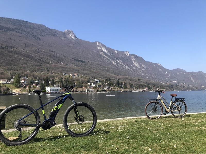 Deux vélos électriques stationnés sur une pelouse au bord d'un lac avec montagnes et ciel bleu en arrière-plan.