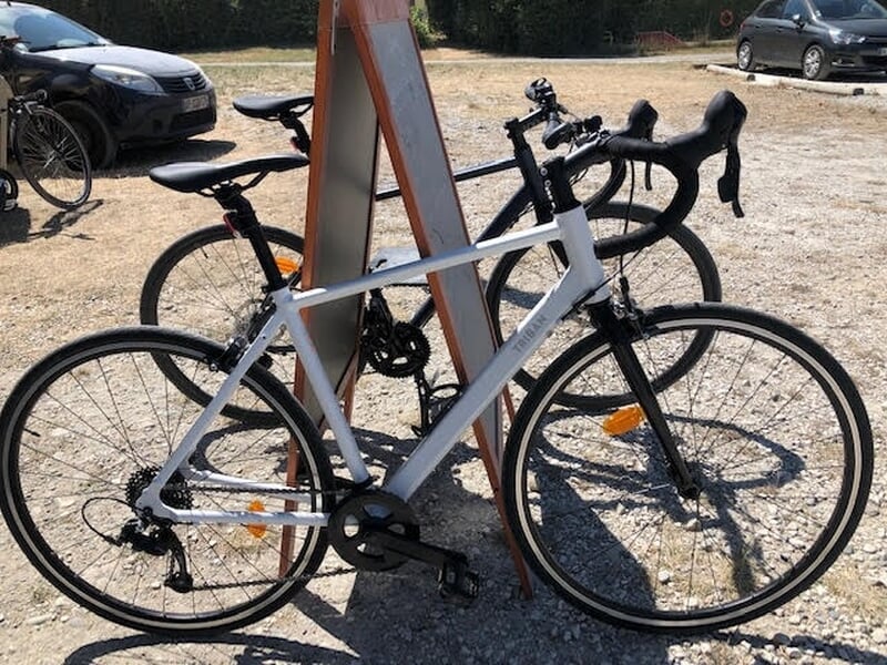 Vélo de route blanc stationné devant un panneau orange avec d'autres vélos et voitures en arrière-plan.