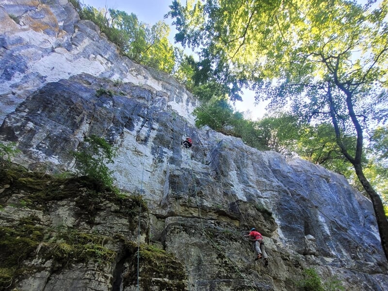 Grimpeur en tenue rouge sur une falaise rocheuse avec vue sur une vallée et montagnes en arrière-plan.
