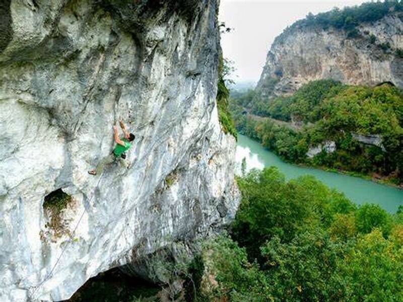 Grimpeur équipé d'un casque et harnais sur une via ferrata rocheuse avec végétation dense autour.
