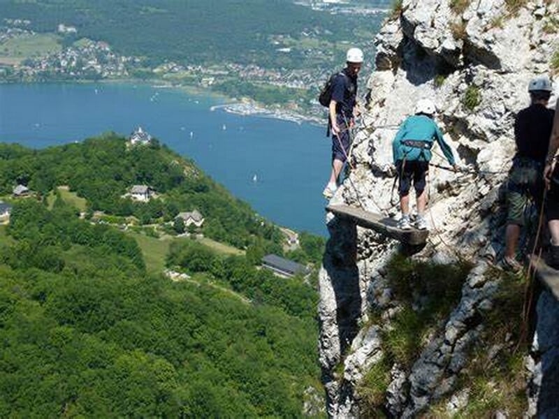Personne en tenue d'escalade sur une via ferrata rocheuse avec arbres verts et ciel bleu en arrière-plan.