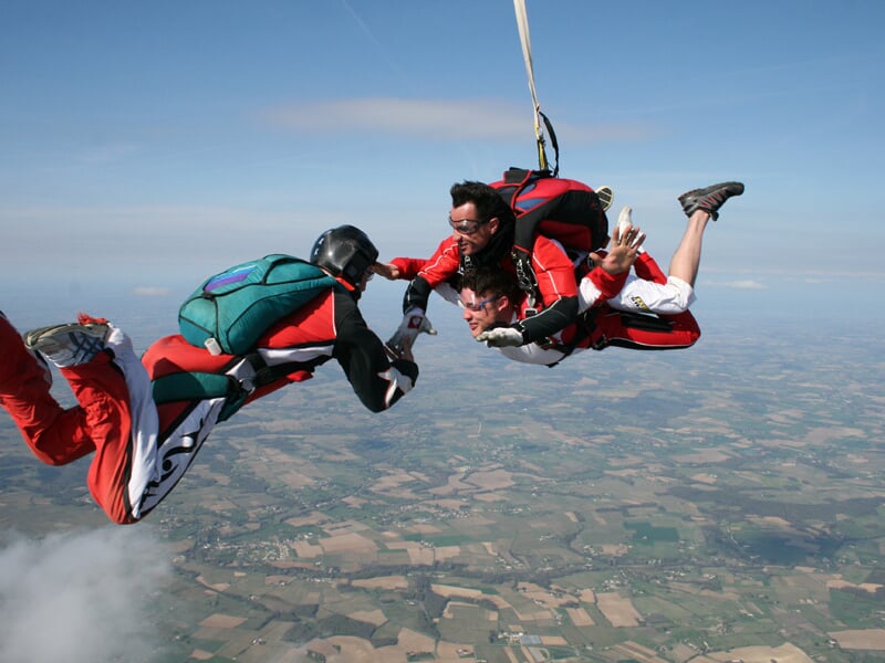 Trois parachutistes en chute libre exécutent une formation aérienne au-dessus d'un paysage agricole. Deux d'entre eux portent des combinaisons rouges et noires tandis que le troisième, équipé d'une co
