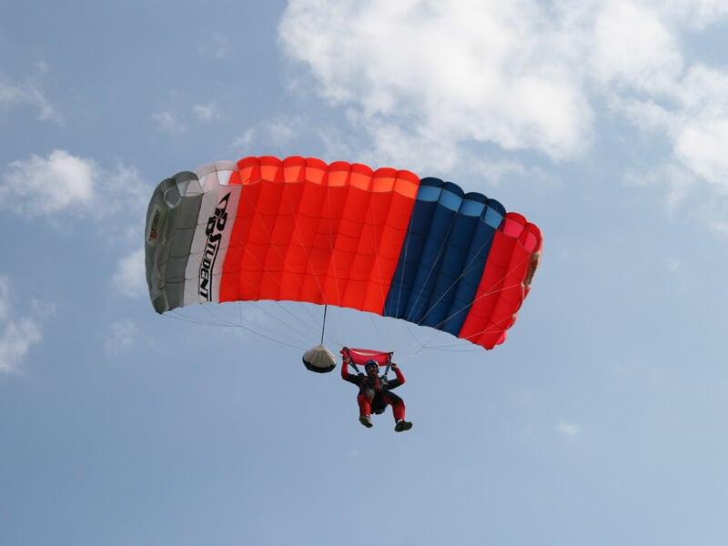 Parachutiste en chute libre avec parachute ouvert rouge, blanc et bleu sous un ciel partiellement nuageux.
