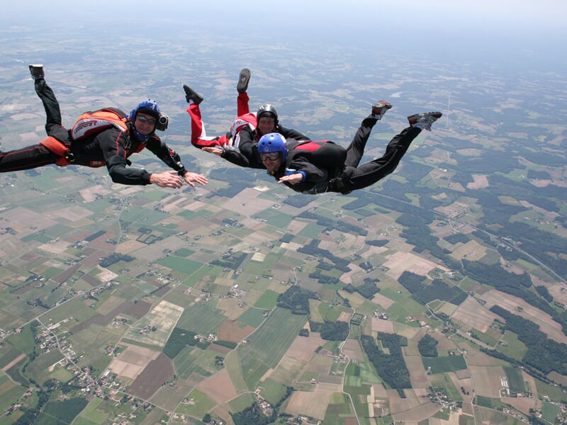 Trois parachutistes en combinaison noire et rouge en formation en chute libre au-dessus d'un paysage rural.