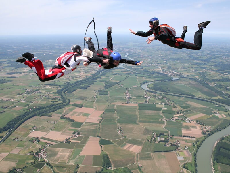 Trois parachutistes en combinaison rouge, blanche et noire en formation en chute libre au-dessus de champs verts.