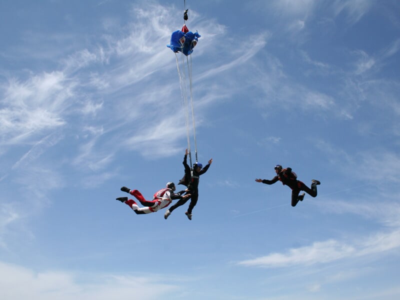 Trois parachutistes en chute libre avec un parachute bleu en train de se déployer dans un ciel clair.