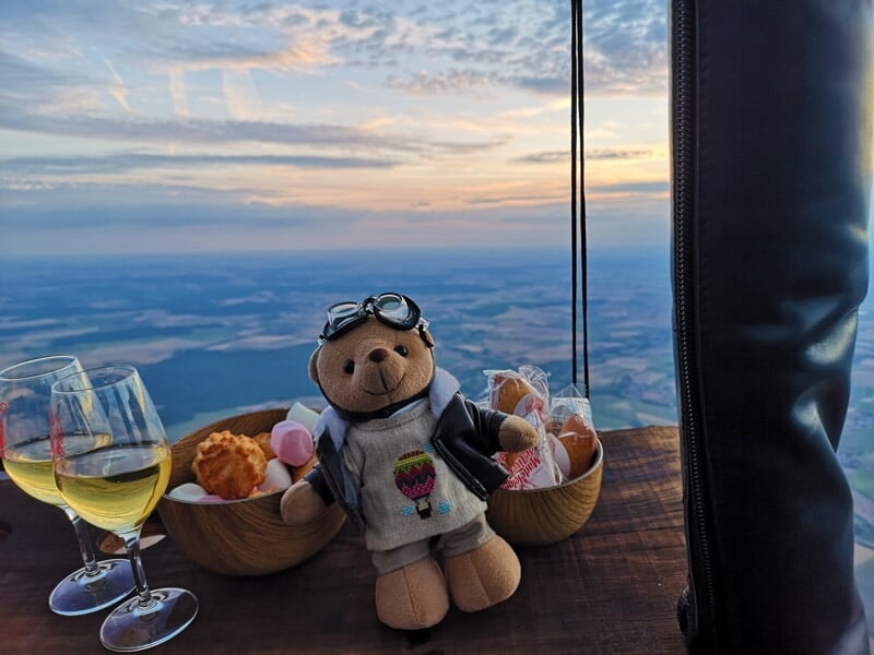 Un ours en peluche dans une montgolfière avec des verres de vin et des pâtisseries au coucher du soleil.
