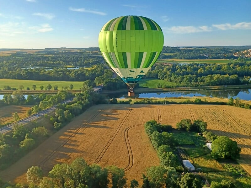 Montgolfière verte volant au-dessus de champs et d'une rivière au coucher du soleil.