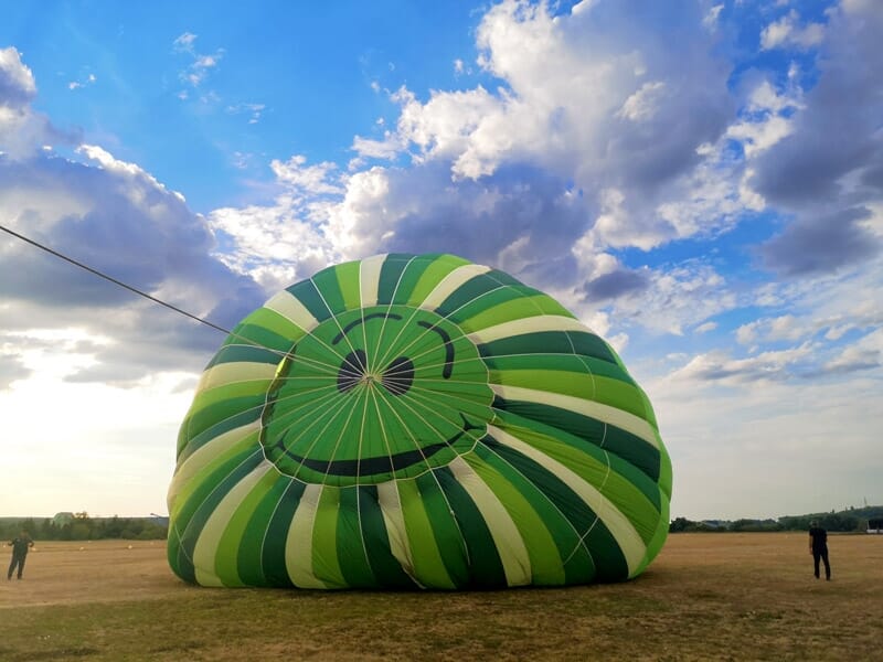 Montgolfière verte et blanche posée au sol, partiellement dégonflée, avec des nuages épais dans le ciel et des silhouettes humaines encadrant la scène.