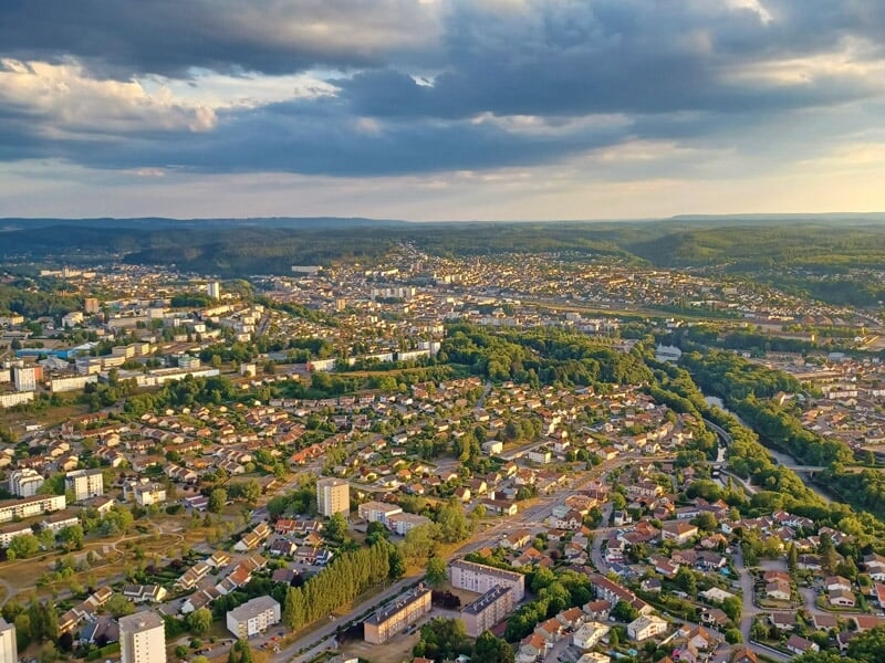 Vue aérienne d’une ville au coucher du soleil, montrant des maisons individuelles, des immeubles et des espaces verts, avec un ciel partiellement nuageux.
