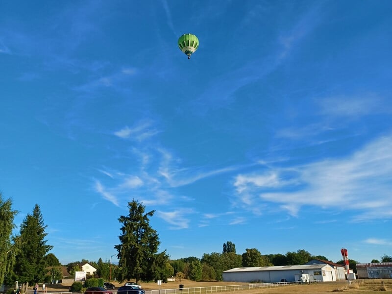 Montgolfière verte en plein vol sous un ciel bleu parsemé de nuages légers, survolant une zone rurale avec des arbres et bâtiments au sol.