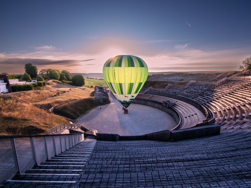 Montgolfière verte volant au-dessus d'un amphithéâtre antique en pierre au soleil.